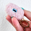 A woman holds up a plush pink AirPod Case Cover with a golden-colored keychain accent and displays its bottom where there is a hole to charge your device - Fleece Chic