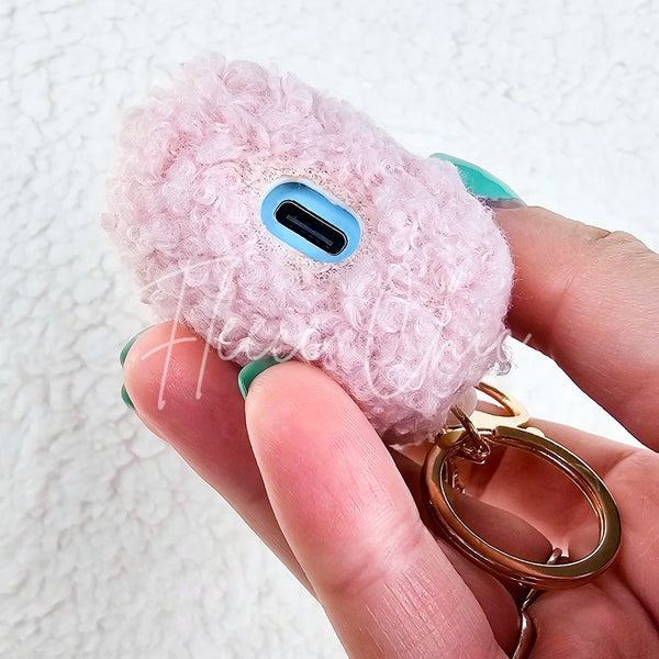 A woman holds up a plush pink AirPod Case Cover with a golden-colored keychain accent and displays its bottom where there is a hole to charge your device - Fleece Chic