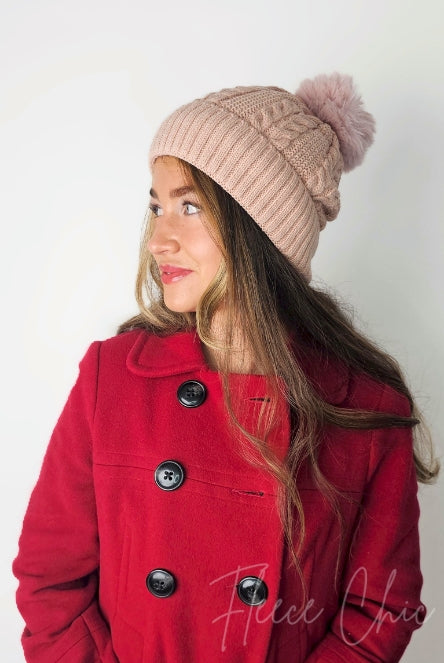 A woman wears a pink fleece beanie with a cute pom pom on it by Fleece Chic paired with a red coat.