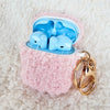 A teddy AirPod Case Cover that is light pink with a golden-colored keychain accent by Fleece Chic and light blue earbuds inside the case.