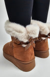 A pair of winter boots that're brown and fur-lined with a back bow are worn by a person in black leggings - Fleece Chic