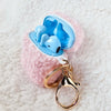 A teddy AirPod Case Cover that is light pink with a golden-colored keychain accent by Fleece Chic withlight blue earbuds inside.