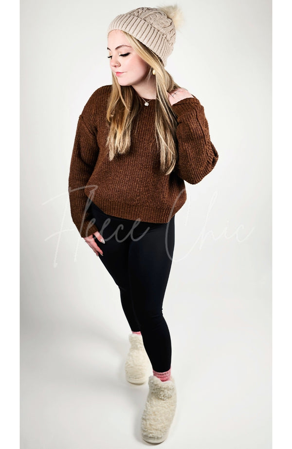 A woman wears Fleece Chic's fleece beanie and sherpa boot slippers paired with black leggings and a brown sweater.