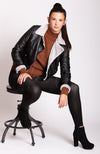 Woman sitting on a stool wearing a black leather jacket with gray fur lining, brown cotton sweater, and black leggings - Fleece Chic