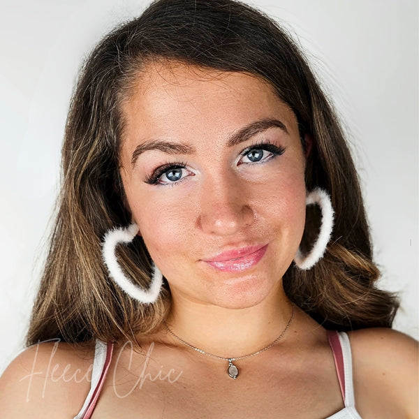A woman wears heart shaped earrings that are white and fluffy by Fleece Chic.
