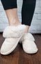 Faux fur slippers in beige with a plush collar are modeled by a woman standing on a dark wood floor - Fleece Chic