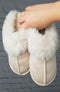 Faux fur slippers in beige with a plush collar are held up infront of a brick background - Fleece Chic